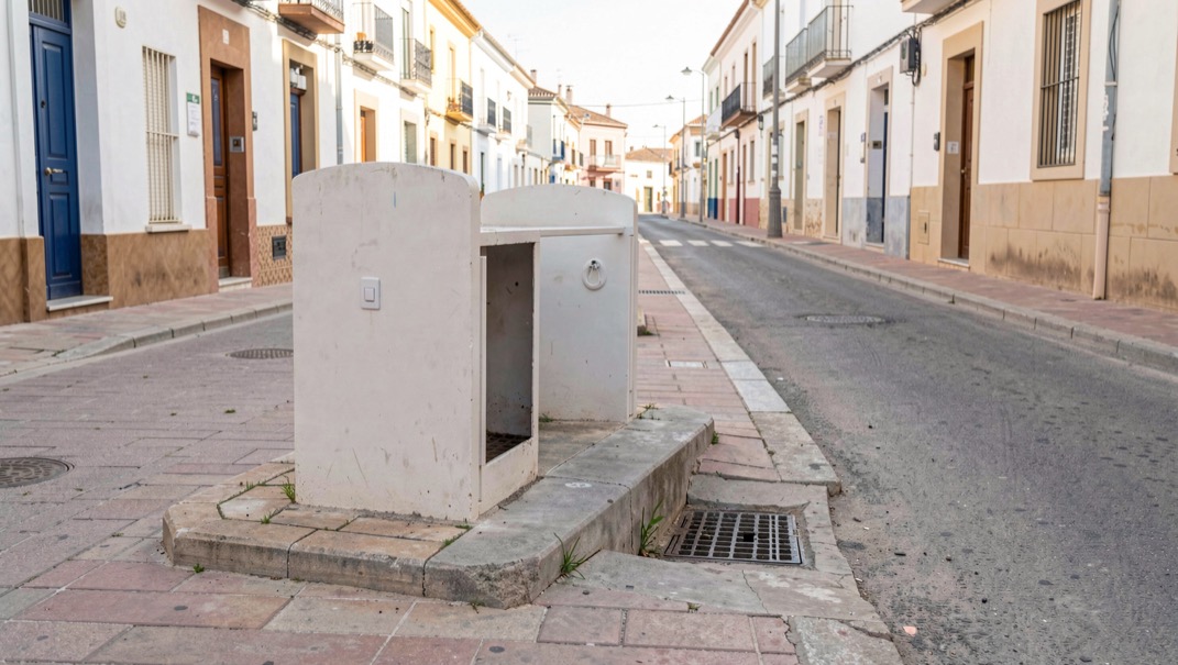 Detalle documental de una arqueta urbana limpia y una conducción de saneamiento en una calle tranquila de Chiclana, luz mediterránea suave, sin personas ni marcas.